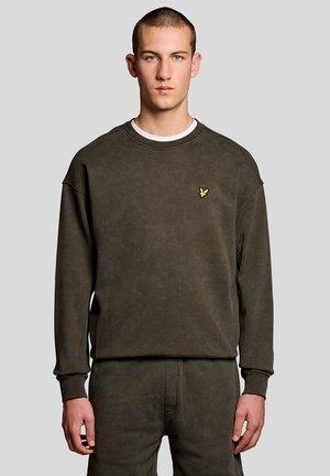 Jeune homme portant un sweat-shirt marron foncé et un pantalon assorti, debout devant un fond gris clair uni.
