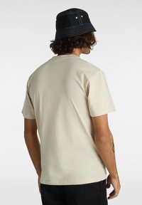 Beige bomullströja med kort ärm, rund halsringning och en slät textur, bärs med en svart bucket hat med kontrasterande vit sömnad.