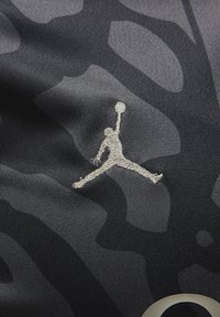 Musta urheiluvaatekangas, jossa on sävyton kamopainatus, sekä näkyvästi brodeerattu Jumpman-logo hopeisena ja ympyräinen korostus kulmassa.