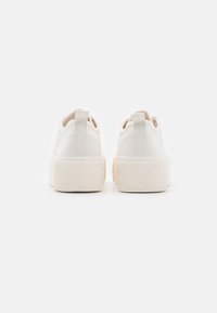 Baskets blanches avec une semelle en caoutchouc texturé, un dessus en toile lisse et un tirant de talon renforcé, présentant un design arrondi à l'arrière.