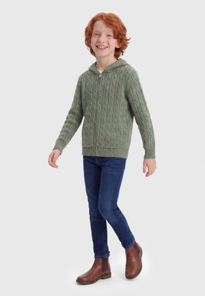 Crvenokosi nasmiješeni dječak stoji, nosi zelenu pletenu majicu s kapuljačom na patentni zatvarač, plave traperice i smeđe kožne gležnjače.