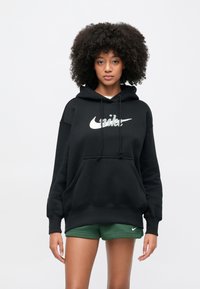 Kvinne med krøllete hår iført en svart Nike-hettegenser og grønne Nike-shorts, stående foran en ensfarget hvit bakgrunn.