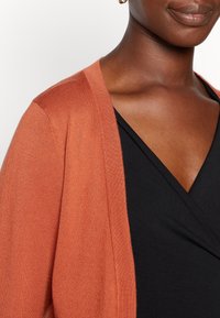 Rostfärgad cardigan med en mjuk, stickad textur, öppen framsida och vida ärmar, bärs över en svart wrap-top. Inga särskilda mönster eller detaljer.