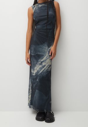 Bezrękawowa długa sukienka w marmurkowy wzór w odcieniach ciemnego niebieskiego i szarości, noszona z czarnymi wsuwanymi butami z ćwiekami, pokazana od szyi do stóp na jednolitym tle.