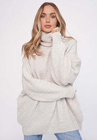 Helles beiges Oversized-Pullover mit hohem Rollkragen, gerippter Textur und weiten Ärmeln, kombiniert mit hellblauen Jeans.