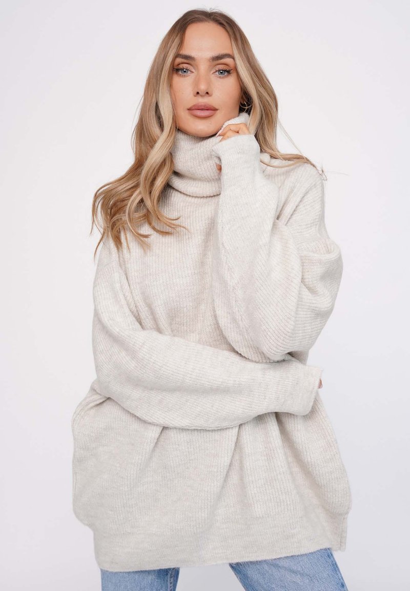 Helles beiges Oversized-Pullover mit hohem Rollkragen, gerippter Textur und weiten Ärmeln, kombiniert mit hellblauen Jeans.