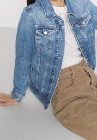 Jeansjacka i ljusblått med spetsig krage, knappstängning, två bröstfickor och långa ärmar. Bärs över en krämfärgad skjorta och beige kordbyxor.