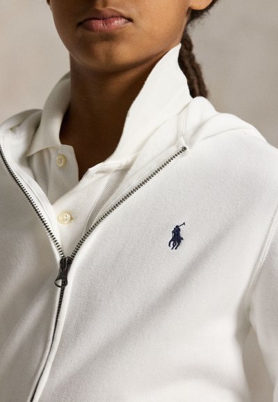 Personne portant une veste blanche zippée par-dessus un polo blanc, avec un logo de joueur de polo bleu marine sur la poitrine de la veste.