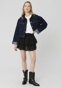 Veste en denim courte avec fermeture à boutons, couleur bleu foncé, associée à une jupe noire à volants superposés et des bottes de cowboy noires.