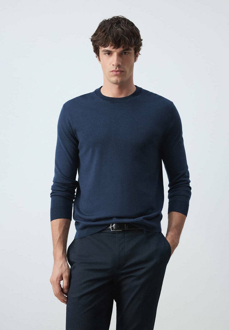 Homme debout de face, portant un pull bleu marine à manches longues et un pantalon foncé, une main dans la poche, sur un fond uni.