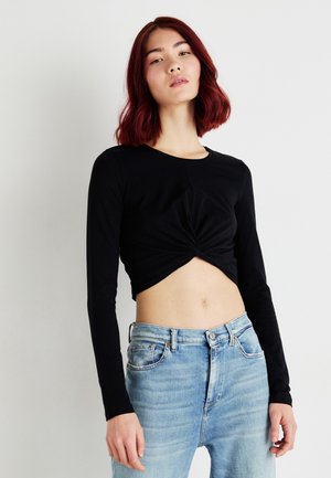 Svart langermet crop-top med vridd frontdetalj, sammen med lyseblå high-waist jeans. Glatt stoff og tettsittende design.