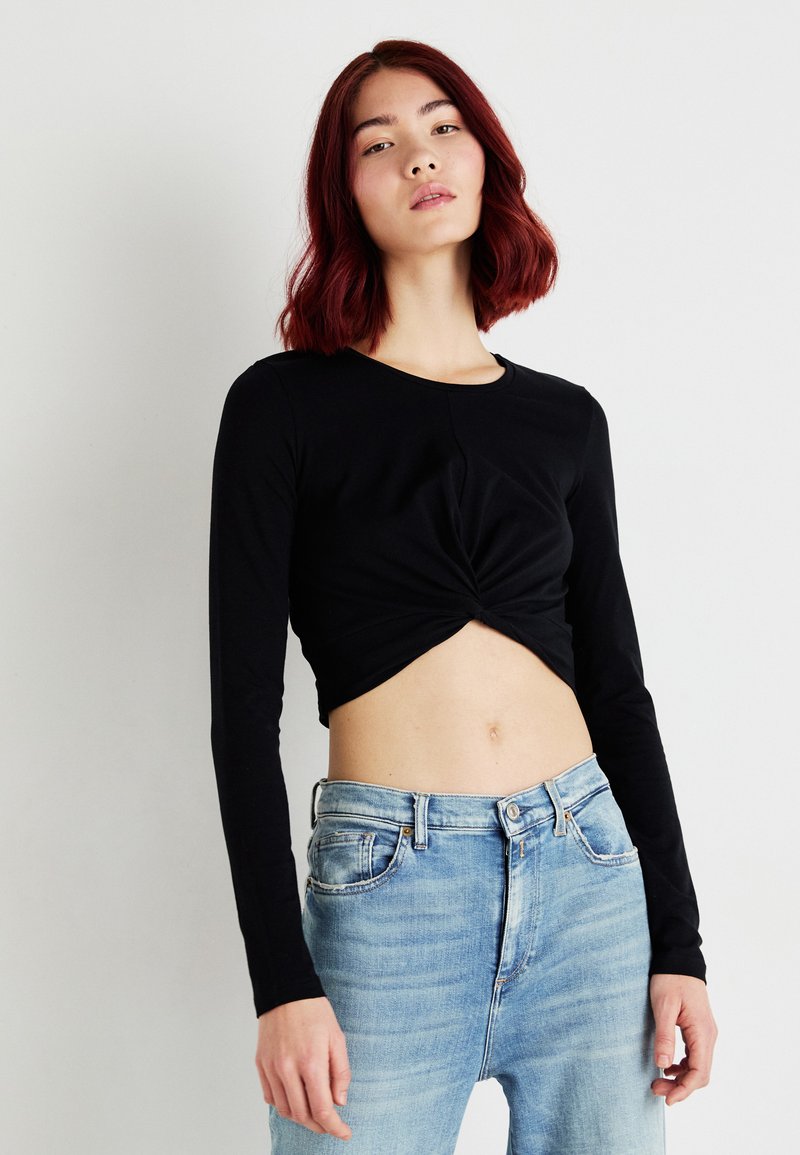 Fekete, hosszú ujjú, cropped top, csavart elülső résszel, világoskék, magas derekú farmernadrággal párosítva. Simább anyag és testhez álló fazon.