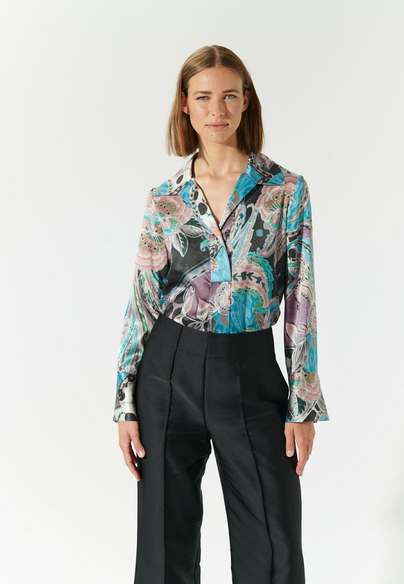 Blusa a fantasia colorata con colletto, caratterizzata da tonalità di turchese, nero e rosa, abbinata a pantaloni neri a vita alta. Texture morbida.