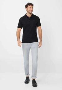 Zwarte polo met korte mouwen, twee knopen bij de kraag, gedragen met lichtgrijze opgerolde jeans en zwarte veterschoenen.