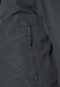 Gros plan sur la poche zippée d'une veste noire avec une languette en tissu sur la fermeture éclair.