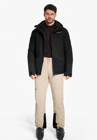 Schwarze isolierte Jacke mit grauen Akzenten, kombiniert mit beigen Schneehosen mit einem dunklen Saum, ergänzt durch schwarze Schneestiefel.