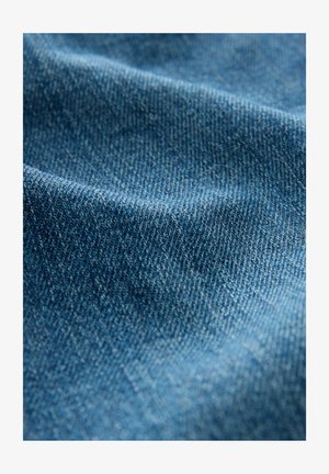 Denima audums ar vidēju zilo krāsu, ar smalku faktūru un diagonālu audumu rakstu, parādot krokas un grumbas.