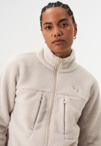 Hellbeige Fleecejacke mit hohem Kragen, Frontreißverschluss und zwei Brusttaschen, aus strukturiertem Stoff mit dezentem Logodetail.