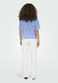 Blusa in pizzo azzurro chiaro con orlo a scallop e maniche a campana, abbinata a pantaloni bianchi, vista da dietro.