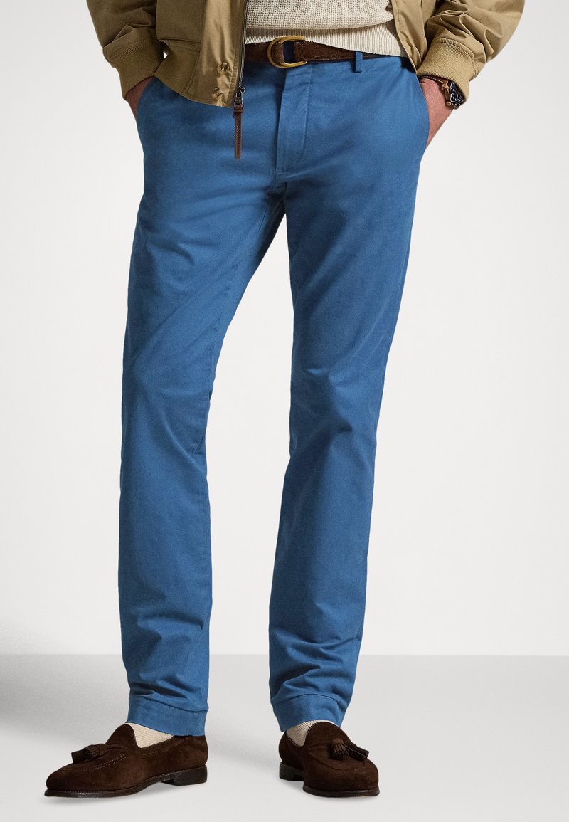 Calças chino de algodão azul com textura suave e corte ajustado, com passadores para cinto e bolsos laterais, combinadas com mocassins castanhos.