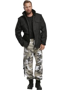 Svart zip-up jacka med två bröstfickor, tillsammans med kamouflage cargo-byxor med sidfickor. Bärs med svarta kängor.