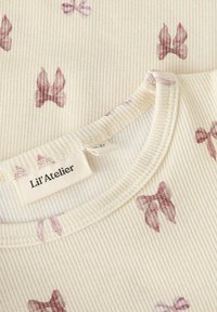 Ribbed cream-colored fabric top featuring a subtle butterfly pattern in mauve tones, with a rounded neckline and a label reading "Li'l Atelier."