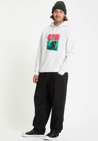 Grijze hoodie met voorop een grafische afbeelding in rood, groen en zwart; zwarte broek met een losse pasvorm; zwarte canvas sneakers; groene gebreide muts.