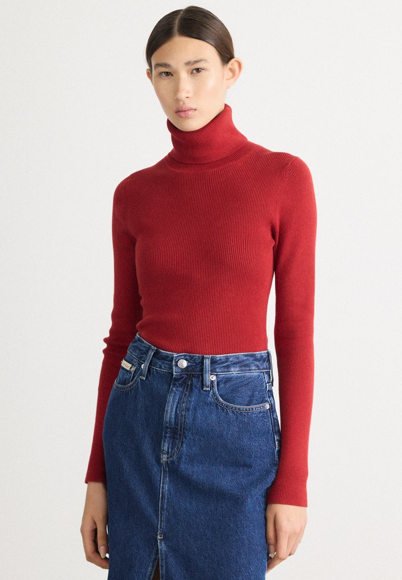 Rdeča rebrasta turtlenecka iz pletenega materiala, kombinirana z jeans krilom, ki ima sprednji razporek in sprednje žepke. Gladka tekstura in oprijet dizajn.