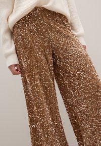 Braune Pailletten-Hosen mit weitem Bein und strukturierter Oberfläche, die ein schimmerndes Finish und eine lockere Passform aufweisen, kombiniert mit einem weißen gerippten Pullover.