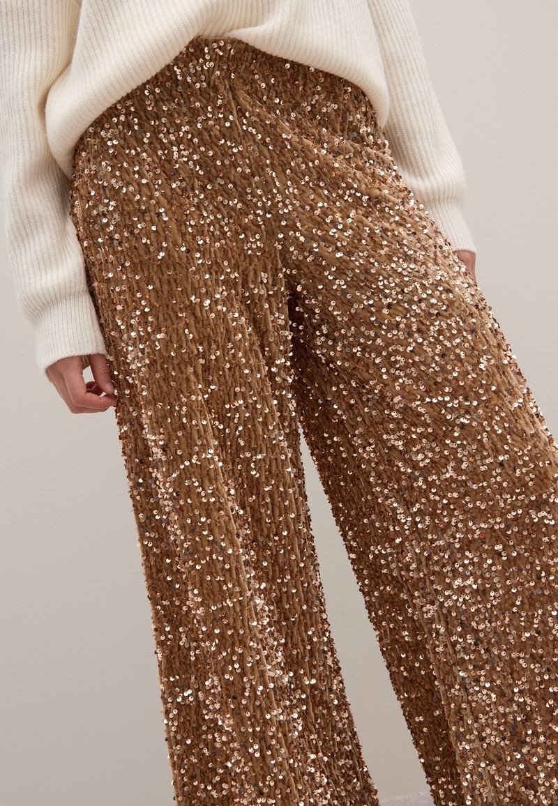 Braune Pailletten-Hosen mit weitem Bein und strukturierter Oberfläche, die ein schimmerndes Finish und eine lockere Passform aufweisen, kombiniert mit einem weißen gerippten Pullover.
