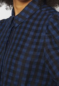 Sininen ja musta ruudullinen paita nappikiinnityksellä, teksturoitu kangas, pehmeä kaulus ja plektit olkapäissä.