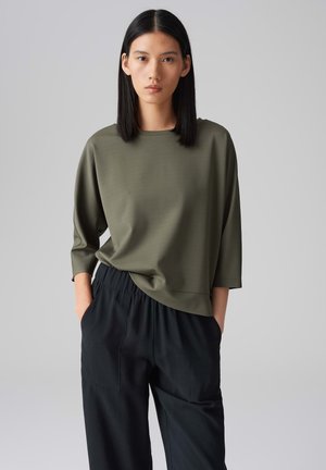 Olivgrünes, locker sitzendes Oberteil mit drei Viertelärmeln, glatter Textur und rundem Ausschnitt, kombiniert mit schwarzen, elastischen Hose mit dehnbarem Bund.