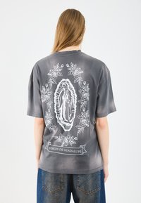 Grafiska pelēka T-kreklā ar balto Pannas de Guadalupe dizainu, ko ieskauj rožu ilustrācijas un baneris, īsas piedurknes, brīva piegriezuma.