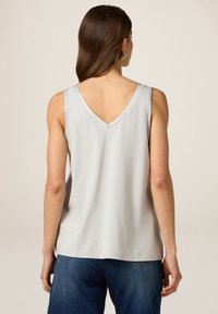 Kvinna i en ärmlös ljusgrå topp med V-formad ryggutskärning och blå jeans, som står mot en neutral bakgrund.