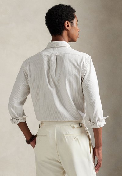 Camisa branca às riscas com mangas arregaçadas, detalhe de prega nas costas; usada com calças de cor clara com bolsos laterais e traseiros.