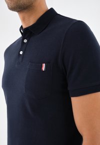 Námořnická modrá polo košile s límečkem, třemi knoflíky a kapsou na levé straně hrudi s červeno-bílo-modrým pruhovaným akcentem.