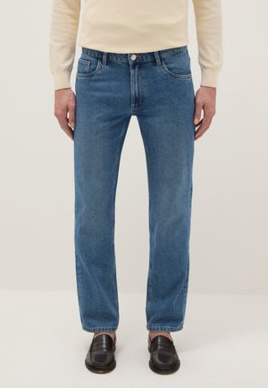 Man draagt een blauwe rechtmodel spijkerbroek, een beige trui en zwarte loafers, staand tegen een effen lichte achtergrond.