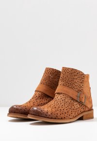 Bottes basses en daim marron avec un motif perforé, bout rond et une bride à boucle. Talon marron uni et semelle texturée complètent le design.