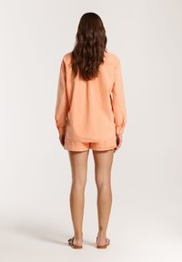Camicia a maniche lunghe color pesca con polsini arricciati, abbinata a pantaloni corti coordinati. Tessuto morbido, vestibilità rilassata e tasche laterali visibili.