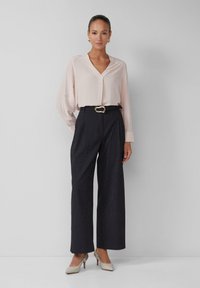 Blouse rose clair à manches longues avec décolleté en V, associée à un pantalon large à rayures sombres et des chaussures à talons hauts crème. Accent ceinture dorée.