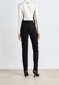 Camiseta blanca de manga larga con acentos negros, cuello alto y logo 'BOSS'. Jeans negros con bolsillos traseros, ajuste slim y etiqueta de cuero marrón. Tacones negros.