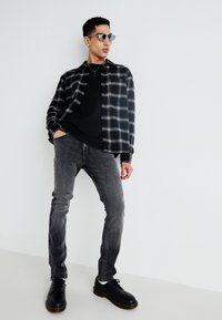 Chemise à carreaux noir et blanc sur un t-shirt noir, jeans gris et bottes noires à lacets avec des accents jaunes. Des lunettes de soleil bleues complètent le look.