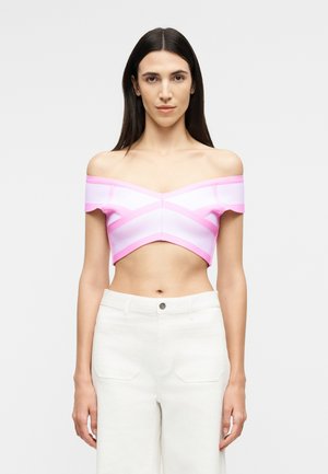 Γυναίκα με μακριά σκούρα μαλλιά, φοράει ροζ και άσπρο ριγέ τοπ που αφήνει τους ώμους ακάλυπτους και λευκό παντελόνι ψηλής μέσης, στέκεται μπροστά σε απλό φόντο.