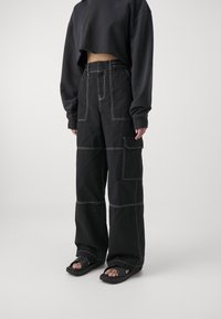 Persoon in zwarte wijde broek met witte stiksels, cropped zwart sweatshirt en zwarte slidesandalen tegen een effen lichte achtergrond.