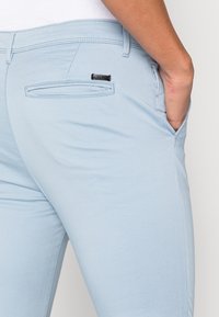 Ljusblå byxor av ett glatt material, med en bakficka med svart accent och en figurnära design.