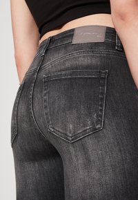 Persona que lleva jeans de mezclilla negros desgastados con costuras visibles en los bolsillos traseros y una etiqueta de cuero gris de la marca en la cinturilla.