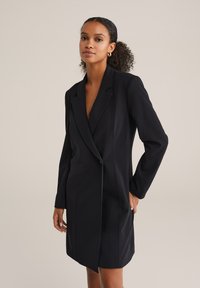 Rochie-jachetă neagră, ajustată, cu mâneci lungi, rever ascuțit și închidere cu un singur nasture. Material neted și tivul până la genunchi.