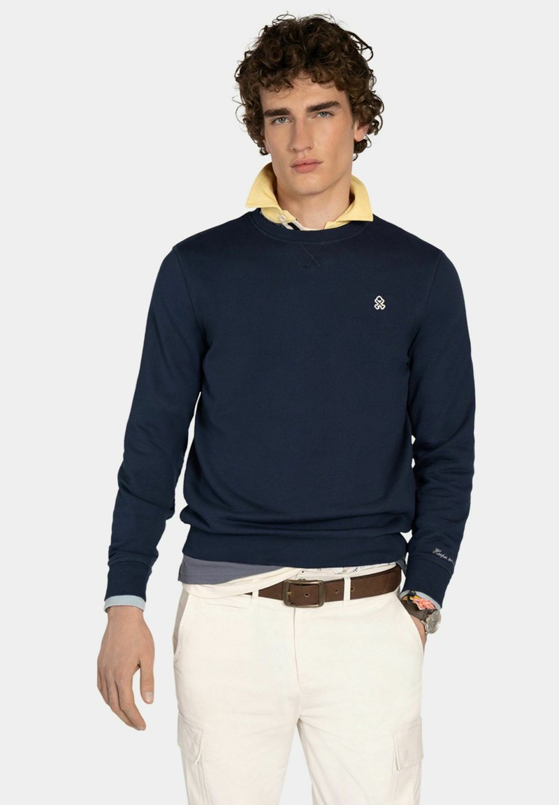 Young man with curly hair wearing navy sweatshirt over yellow collared shirt, white pants, brown belt, and watch, posing against plain background.