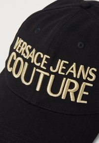 Czarna czapka baseballowa z złotym haftem, na którym widnieje napis "VERSACE JEANS COUTURE". Napis ma teksturowane wykończenie w zestawieniu z gładkim materiałem.