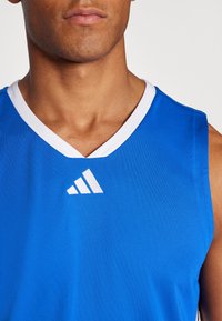 Kék sportos trikó fehér V-nyakkal és kontrasztos fehér logóval, amely három csíkkal rendelkezik, légáteresztő anyagból készült.